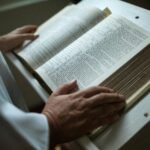 A person in white garment holds an open religious scripture, focusing on hands and text.